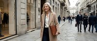 Confident blonde businesswoman walking through historic European city street wearing beige coat, smiling while carrying leather bag during busy day.