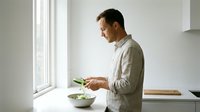 Young Caucasian man in casual shirt using smartphone while eating healthy salad in modern minimalist kitchen by window during morning routine.
