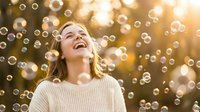 Young Caucasian woman laughing joyfully surrounded by floating soap bubbles in warm golden sunlight creating magical outdoor portrait for lifestyle content.