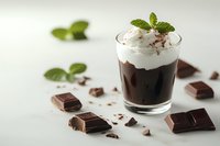 Chocolate dessert with whipped cream and mint garnish in glass, surrounded by dark chocolate pieces on light surface. Fresh mint leaves add elegant touch.