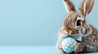 Adorable brown bunny holding blue polka dot Easter egg against light blue background. Festive spring holiday composition with copy space.