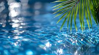 Tranquil swimming pool water surface with sunlight reflections and palm leaf, creating rippling patterns and bokeh effects in vibrant blue tones.