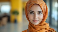 Young woman wearing orange hijab smiling at camera with gentle expression against blurred modern interior background, representing diversity and inclusion.