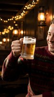 Man in burgundy striped sweater holding beer mug, smiling in warm-lit pub setting with string lights and lanterns. Perfect for beverage, hospitality, celebration, and lifestyle marketing.