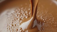 Liquid chocolate stream pouring into melted chocolate surface with splashes and drops creating ripples, macro shot with soft focus and bokeh effect.