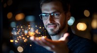 Bearded Caucasian man in glasses holding magical glowing particles in hands against dark background with bokeh lights, expressing wonder and imagination.
