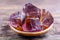 Raw purple fluorite crystals in wooden bowl on rustic table, natural mineral specimen with translucent structure and deep violet color showing crystalline formation.