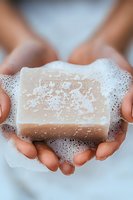 Hands holding natural handmade soap bar with foam bubbles against light background, demonstrating eco-friendly skincare and personal hygiene products.