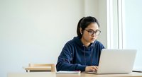 Young South Asian woman wearing glasses and headphones, working on laptop at desk with books, suitable for remote work, education, online learning, and professional development concepts.