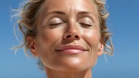 Mature woman with closed eyes enjoying sunshine against clear blue sky, expressing serenity and mindfulness in outdoor wellness moment.