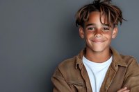 Young mixed race teen with trendy hairstyle and confident smile wearing brown jacket and white t-shirt against gray background, arms crossed in casual pose.