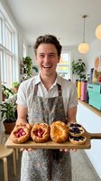 Young male baker with light skin tone and dark hair displaying freshly baked fruit tarts and pastries on wooden board in bright, modern kitchen with plants and pendant lighting.