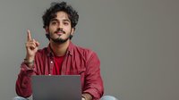 Young South Asian man with curly hair raising index finger in eureka moment while working on laptop against neutral gray background.