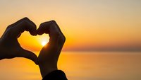Hands forming heart shape silhouette against golden sunset over calm water. Romantic love symbol gesture at peaceful evening seascape for Valentine's Day concepts.