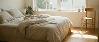 Serene bedroom with unmade bed, natural linen bedding, wooden chair, potted plant, and bright sunlight streaming through windows for home decor and interior design projects.