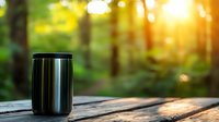 Stainless steel travel mug on wooden table outdoors with blurred forest and golden sunlight in background, morning atmosphere creates peaceful mood.