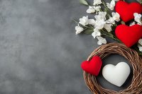 Romantic composition with red and white plush hearts in woven wreath nest, decorated with delicate white flowers against dark gray textured background. Copy space.