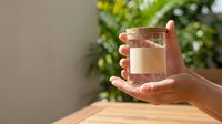 Person with medium skin tone holding a clear glass of filtered water on wooden surface with green foliage background, suitable for health, wellness, hydration and lifestyle content.