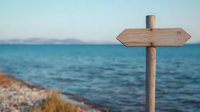 Wooden directional signpost on coastal beach with blue ocean and distant mountains, perfect for travel and navigation concepts.