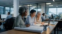 Diverse female architects and engineers reviewing blueprints and laptop in modern office workspace, collaborating on construction project planning and design development.