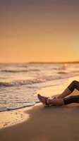 Woman in athletic leggings and sneakers resting on sandy beach at golden sunset, waves gently washing ashore in warm evening light.