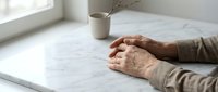 Elderly hands resting on white marble table surface in bright minimalist interior with ceramic vase and dried branches creating peaceful contemplative atmosphere.