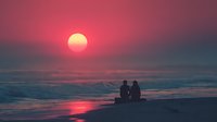 Romantic couple silhouettes sitting on beach watching stunning sunset over ocean waves with pink and purple sky colors reflecting on water surface.