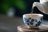 Pouring hot tea from white teapot into traditional Asian ceramic cup with blue floral pattern on wooden surface against dark green background. Cozy drink moment.