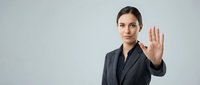 Professional businesswoman in dark suit making stop gesture with raised hand against neutral background for corporate communication concepts.