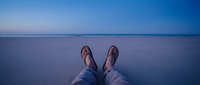 Young adult male wearing casual sandals and blue jeans sitting at edge of calm ocean water at dusk, feet dangling over serene seascape horizon.