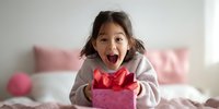 Excited young Asian girl with surprised expression holding pink gift box with red bow on bed, expressing joy during celebration moment.