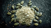 Raw pumpkin seeds scattered around pile of ground seed flour powder on dark stone background, macro food photography for healthy eating concept.