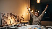 Young Asian woman celebrating success in creative workspace with mood boards and design materials, arms raised in victory gesture at night.