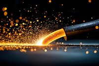 Glowing hot metal sparks scatter dramatically from welding torch against dark background, creating mesmerizing pattern of golden light particles in motion.