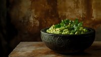 Fresh homemade guacamole with cilantro garnish served in rustic stone bowl against dark moody background on wooden surface. Perfect for food blogs.