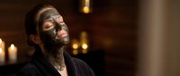 Young Caucasian woman in robe relaxing with dark clay mud mask on face at luxury spa, candles glowing in background.