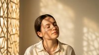 Young adult male with East Asian ethnicity, wearing a light beige linen shirt, eyes closed in contemplation, bathed in warm golden sunlight filtering through geometric window patterns, serene indoor portrait.