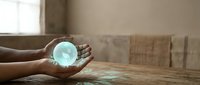 Hands holding glowing turquoise orb of light in warm sunlit room with wooden table and cardboard boxes creating magical energy concept for meditation.