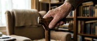 Elderly man's weathered hand gripping wooden walking cane in cozy home library setting with bookshelves and comfortable furniture in background.