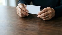 Senior man with medium skin tone holding blank white card at wooden table, wearing dark business attire for corporate messaging and branding applications.