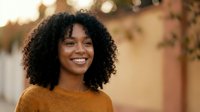 Happy young African American woman with curly hair smiling outdoors in warm autumn light wearing cozy orange sweater for lifestyle and portrait concepts.