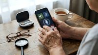 Senior person using voice assistant app on smartphone with wireless earbuds and coffee cup on wooden table for hands-free communication and accessibility.