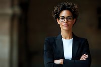 Young African American businesswoman wearing glasses and black blazer with confident pose and crossed arms, professional corporate portrait against dark background.