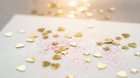 Gold heart and star confetti scattered on white surface with pink glitter, soft bokeh lights in background, festive romantic celebration.