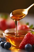 Golden honey dripping from wooden spoon into glass bowl with fresh strawberries and blueberries, macro food photography showing liquid texture and natural sweetness.