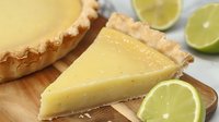 Fresh key lime pie slice on wooden board with citrus garnish, creamy yellow filling in flaky golden crust, bright and zesty dessert presentation.