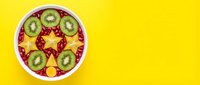 Colorful fruit bowl with kiwi slices, berries, and tropical fruits arranged in star pattern on bright yellow background, overhead view for food and wellness content.