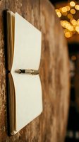 Open notebook with blank pages attached to rustic wooden wall with metal clip, warm bokeh lights in background, suitable for journaling, writing, education, and creative projects.
