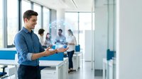 Young Caucasian businessman using tablet in modern office with team meeting in background, corporate technology and workplace collaboration.