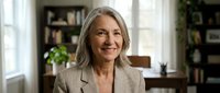 Mature woman with long gray hair wearing beige blazer, smiling in home office with bookshelves and plants, suitable for business, corporate, and lifestyle content.